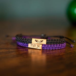 Op de afbeelding: Twee macram&eacute; armbanden, &eacute;&eacute;n zwart en &eacute;&eacute;n paars, zijn op een houten oppervlak gestapeld. Elke armband heeft een rechthoekig houten accentstuk met een vleugelontwerp en de datum "11.07.24" gegraveerd. De armbanden zijn verstelbaar.