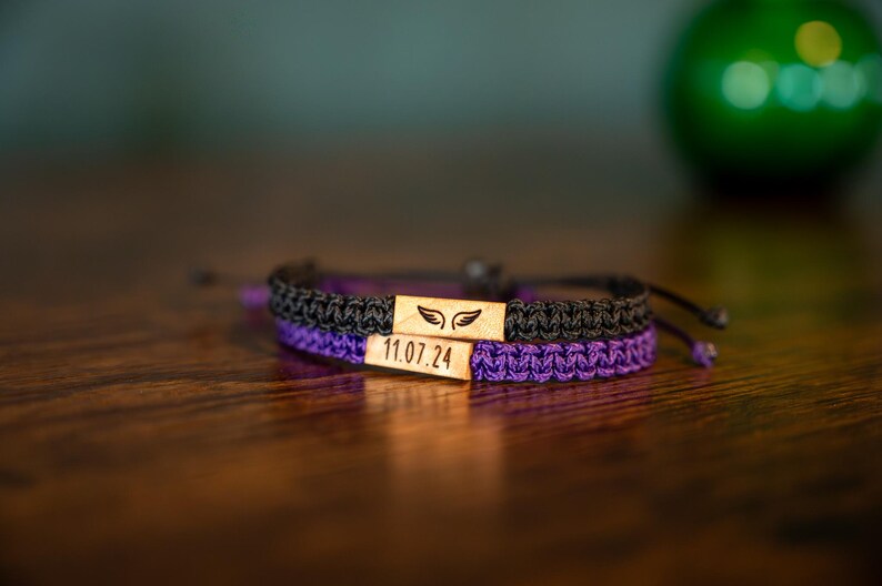 Op de afbeelding: Twee macram&eacute; armbanden, &eacute;&eacute;n zwart en &eacute;&eacute;n paars, zijn op een houten oppervlak gestapeld. Elke armband heeft een rechthoekig houten accentstuk met een vleugelontwerp en de datum "11.07.24" gegraveerd. De armbanden zijn verstelbaar.