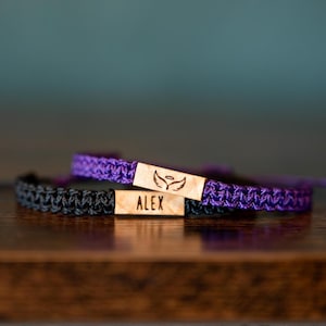 May include: Two macrame bracelets, one black and one purple, with wooden nameplates. The black bracelet has a wooden plate with the name "ALEX" engraved. The purple bracelet has a wooden plate with an angel wing design.