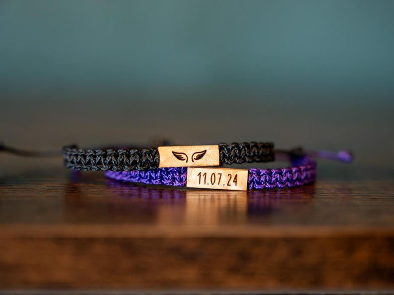 Op de afbeelding: Twee macram&eacute; armbanden, &eacute;&eacute;n zwart en &eacute;&eacute;n paars, met rechthoekige houten accenten. De zwarte armband heeft een vleugelontwerp en de paarse armband heeft de datum "11.07.24" gegraveerd.