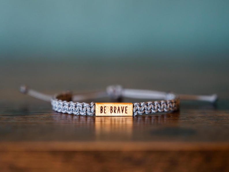 May include: A gray braided bracelet with a wooden rectangular accent piece. The accent piece has the words "BE BRAVE" engraved on it. The bracelet is adjustable and sits on a dark brown wooden surface.
