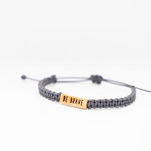 May include: Gray macrame bracelet with a wooden rectangular bead that reads "BE BRAVE". The bracelet is adjustable with sliding knots and is displayed on a white background. A simple, inspirational accessory.