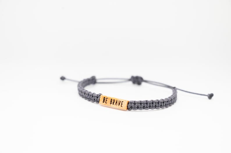 May include: Gray macrame bracelet with a wooden rectangular bead that reads "BE BRAVE". The bracelet is adjustable with sliding knots and is displayed on a white background. A simple, inspirational accessory.
