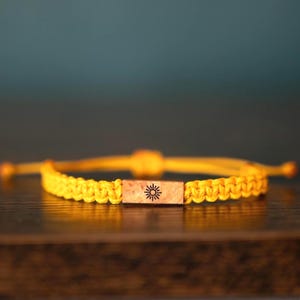 May include: A yellow cord bracelet featuring a rectangular wooden centerpiece. The wooden piece has a natural light brown grain and is engraved with a small black sun icon. The bracelet is constructed with a braided cord design and adjustable ends.