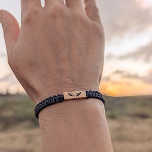 Op de afbeelding: Zwart gevlochten armband met een rechthoekig houten middelpunt. Het middelpunt heeft een vleugelontwerp. De armband wordt om een pols gedragen tegen een vage achtergrond van een zonsondergang.