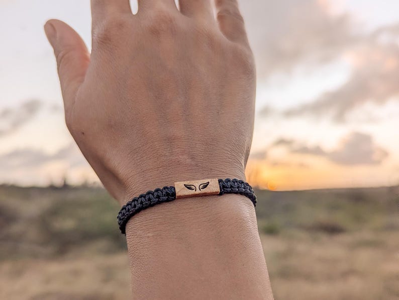 Op de afbeelding: Zwart gevlochten armband met een rechthoekig houten middelpunt. Het middelpunt heeft een vleugelontwerp. De armband wordt om een pols gedragen tegen een vage achtergrond van een zonsondergang.