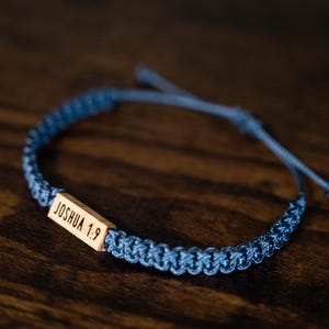 May include: A blue braided cord bracelet featuring a rectangular wooden bead engraved with the text JOSHUA 1:9. The bracelet has an adjustable sliding knot closure and a woven texture throughout the band, showcasing a natural wood finish.