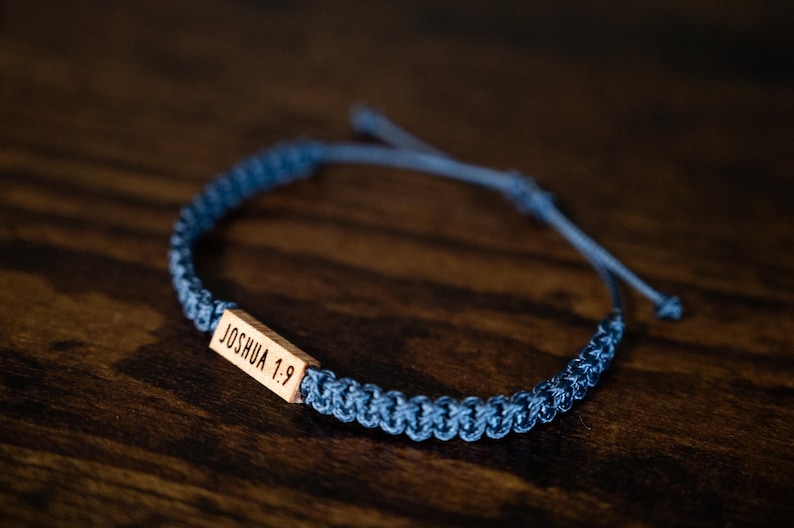 May include: A blue braided cord bracelet featuring a rectangular wooden bead engraved with the text JOSHUA 1:9. The bracelet has an adjustable sliding knot closure and a woven texture throughout the band, showcasing a natural wood finish.
