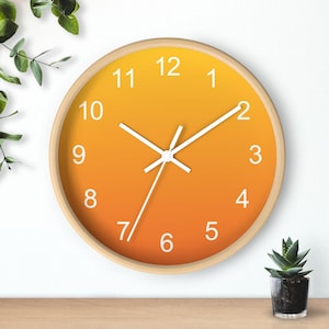 May include: Round wall clock with a gradient orange face and white hands and numerals. The clock has a light wood frame and is set against a white wall. The clock is a decorative item.