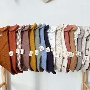 Peut inclure: Une présentation de bavoirs pour bébés de différentes couleurs et motifs, y compris des teintes unies et du vichy. Les bavoirs sont suspendus à un portant en bois, chacun avec une petite étiquette. Un bavoir de couleur neutre est visible en arrière-plan.