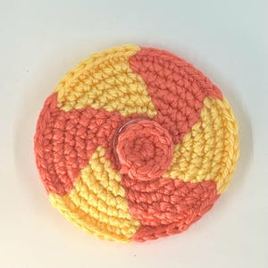 May include: A crocheted coaster with a yellow and coral swirl pattern. The coaster is round and has a small, crocheted circle in the centre.
