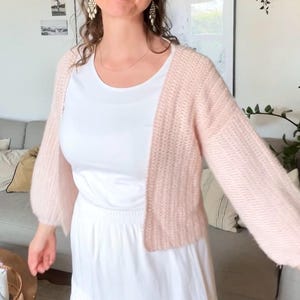 Könnte beinhalten: Eine hellrosa, vorne offene Strickjacke mit langen, leicht bauschigen Ärmeln und einem strukturierten Rippenmuster. Die Strickjacke wird über einem schlichten weißen T-Shirt mit kurzen Ärmeln und einem langen, weißen Stufenrock mit Gummizug getragen.