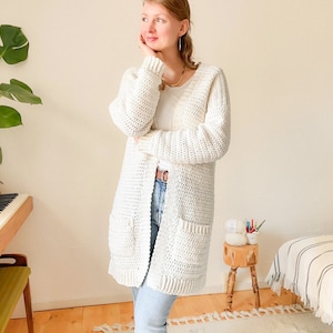 Op de afbeelding: Een wit gebreid vest met lange mouwen en twee zakken. Het vest wordt gedragen over een wit T-shirt en een blauwe jeans.