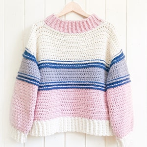 beautiful multicolor sweater hanging on  white wooden wall. Crochet sweater is light pink, soft white, dark blue stripes, light blue middle section, light pink stomach area with soft white finish & light pink neckline finish. Crochet sweater pattern.