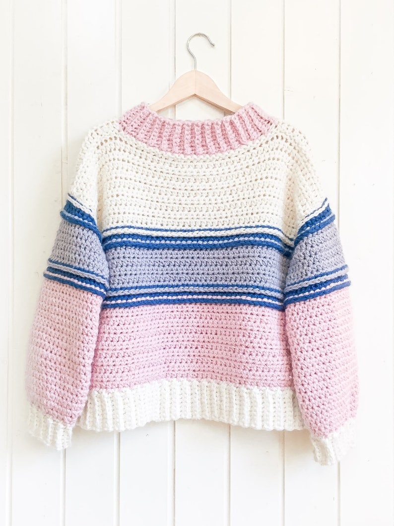 beautiful multicolor sweater hanging on  white wooden wall. Crochet sweater is light pink, soft white, dark blue stripes, light blue middle section, light pink stomach area with soft white finish & light pink neckline finish. Crochet sweater pattern.