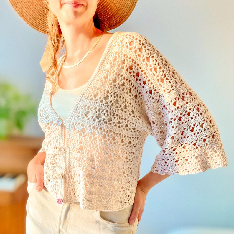May include: A white crochet cardigan with a V-neck and 3/4 sleeves. The cardigan has a delicate openwork pattern and is made with a soft yarn.