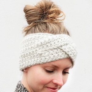 May include: A white crocheted headband with a twisted design. The headband is worn by a person with brown hair pulled back in a bun.