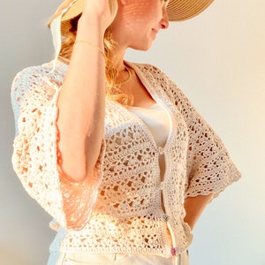 May include: A white crochet top with a button closure and bell sleeves. The top is worn over a white tank top and paired with white denim shorts. The model is wearing a straw hat with a white ribbon.