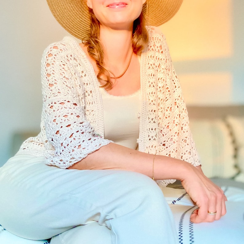 May include: A woman wearing a white crocheted cardigan with a straw hat and white pants.