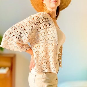 May include: A crocheted cardigan with a delicate openwork pattern in a cream color. The cardigan has a relaxed fit and three-quarter sleeves.