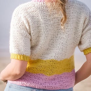 Puede incluir: Un top de ganchillo color crema con mangas cortas y un diseño de bloques de color. El top presenta una franja amarilla y rosa en la sección media y un escote rosa. La prenda se combina con unos vaqueros azules.