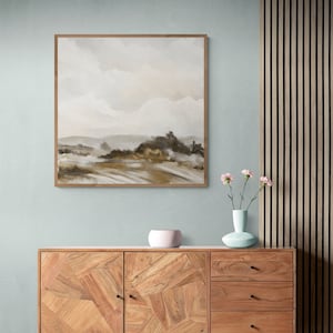 May include: A framed abstract landscape painting with muted brown, beige, and white colors. The painting depicts a hazy, distant landscape with hills and clouds. The painting is hanging on a light blue wall above a wooden cabinet.