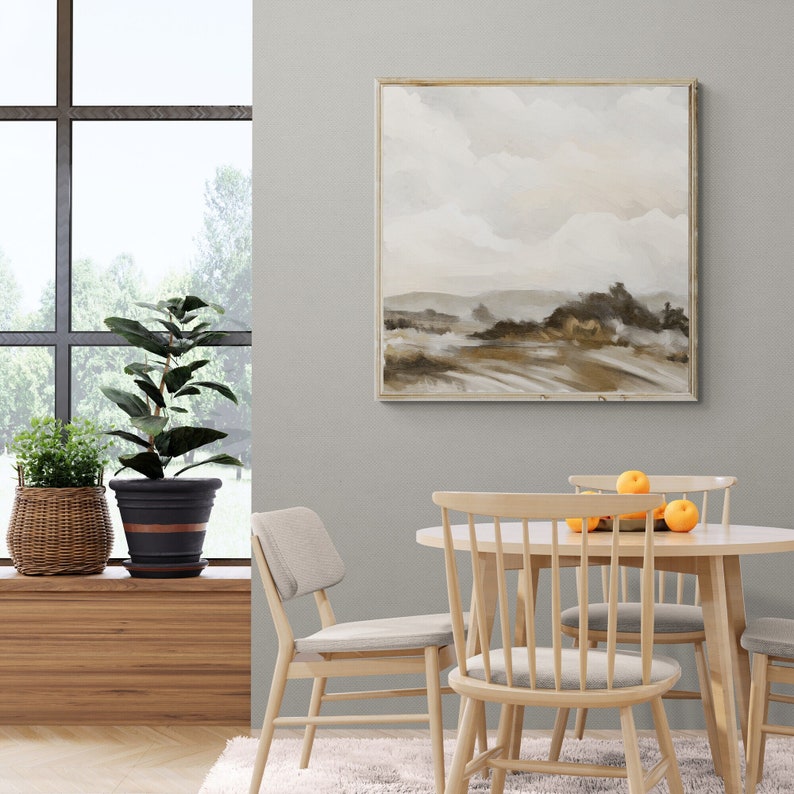 May include: A light brown abstract landscape painting in a light wood frame hangs on a light gray wall above a wooden table with four light wood chairs. A potted plant sits on a windowsill to the left of the table.