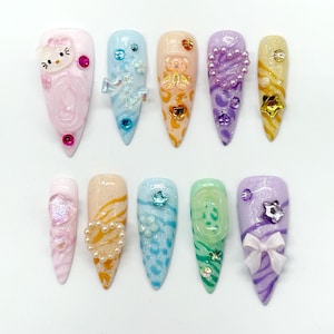 Könnte beinhalten: Ein Set aus zehn mandelförmigen Press-on-Nägeln in Pastellfarben wie Rosa, Blau, Pfirsich, Lila, Gelb und Grün. Die Nägel sind mit 3D-Anhängern verziert, darunter ein Katzengesicht, ein Teddybär, Schleifen, Herzen, Sterne und Perlen.