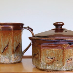 May include: A brown ceramic jug and a lidded container. The jug has a handle and a spout. The container is square with a lid and a small knob. Both pieces have a textured, earthy design with brown, tan, and green hues.