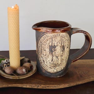 May include: A tall, cylindrical beeswax candle with a textured honeycomb pattern sits in a small, round ceramic dish alongside a dark brown ceramic pitcher. The pitcher features a large, circular embossed medallion with intricate medieval-style imagery.