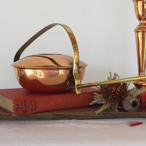 May include: A polished copper bowl with a lid and a long brass handle, resting on a red book titled Dot and the Kangaroo. Beside it stands a tall copper candlestick, a dried flower, and a wooden board with a natural, irregular edge.