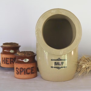 May include: A large beige ceramic salt pig with a wide opening and the word SALT printed on the front, accompanied by two smaller brown ceramic jars with cork lids labelled HERB and SPICE, and a small bundle of dried wheat stalks.