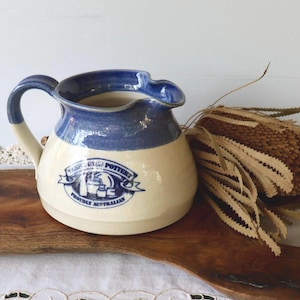 May include: Cream and blue pottery jug with a handle and spout. The jug has a blue rim and features the "Tarragindi Pottery Proudly Australian" logo. It is displayed on a wooden board, with dried foliage.