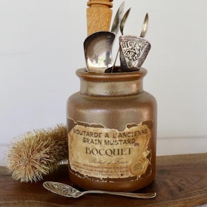 May include: A brown ceramic mustard jar labelled MOUTARDE A L'ANCIENNE GRAIN MUSTARD BOCQUET with a torn paper label. Inside are several silver spoons and a wooden honey dipper. A small round brush and a silver spoon lie in front of the jar.