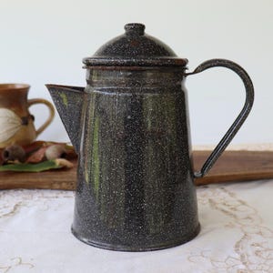 May include: A vintage black speckled enamel coffee pot with a lid and handle. The coffee pot has a spout for pouring and is on a white lace tablecloth. A brown mug is in the background.