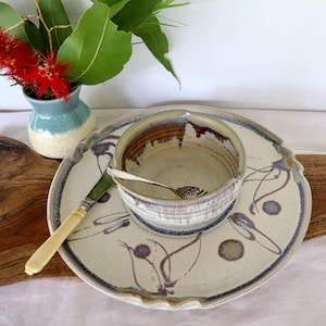 May include: A ceramic bowl and matching plate set featuring a cream-coloured glaze with hand-painted purple and brown floral motifs. A silver spoon rests inside the bowl, and a knife with a cream handle lies on the plate. The edges are scalloped.