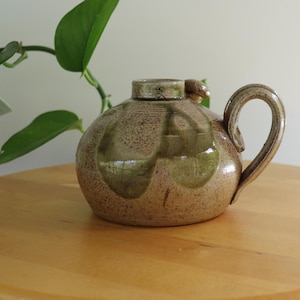 May include: A handmade ceramic jug with a rounded form and a curved handle. The jug has a speckled brown glaze with green leaf-like designs. It rests on a wooden surface, with a plant and another vase in the background.
