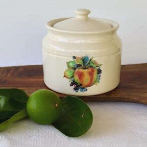 May include: A cream-coloured ceramic jar with a lid, decorated with a colourful fruit illustration. The jar has a round shape and a small handle on the lid. A lime and green leaves are in the foreground.