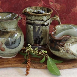 May include: Three handcrafted ceramic jugs in shades of green, brown, and black. The jugs have unique shapes and designs, including handles and spouts. They are displayed on a wooden surface with a sprig of greenery.