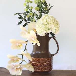 May include: A brown ceramic jug with a handle, holding a floral arrangement of white and green flowers and pale yellow orchids. The jug features a geometric design at the base and rests on a wooden surface.