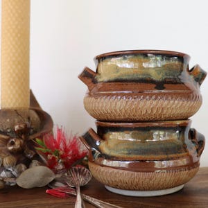 May include: Two stacked ceramic bowls with handles, featuring a glossy brown and tan glaze on the upper portion and a textured, light brown base. The bowls have a rounded shape and are positioned next to a tall candle and a small metal spoon.
