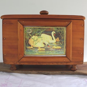 May include: A rectangular wooden box with a hinged lid and small, rounded feet. The front has a framed image of swans in a pond with the text "Butter Scotch Callard & Bowser". The box is made of light brown wood.