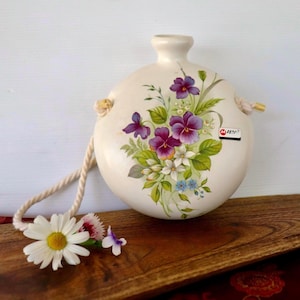 May include: A round, cream-coloured ceramic canteen featuring a hand-painted floral design of purple pansies, white blossoms, and green leaves. The vessel has a narrow neck, a thick beige rope handle, and a small rectangular label with the text M ZERA.