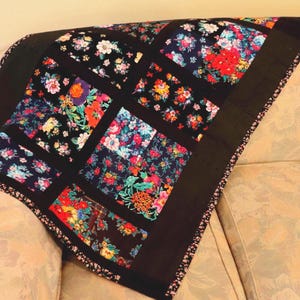 May include: A floral quilt with a black border and backing, draped over a sofa. The quilt features square panels of colorful flowers on a black background. The flowers are in shades of red, pink, yellow, and blue.