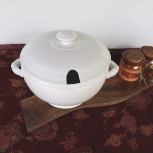 May include: A white ceramic soup tureen with a lid and a ladle opening. The tureen has two handles and sits on a wooden serving board. Two small jars with cork lids are visible in the background. The background is a patterned red tablecloth.