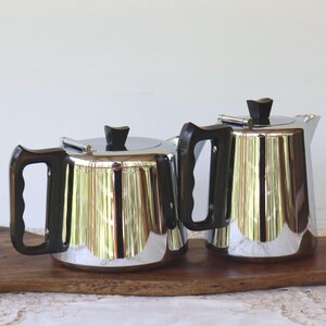 May include: Two polished chrome teapots with black handles and lids. They sit on a wooden tray, reflecting the light. The teapots have a classic, streamlined design, suggesting a vintage aesthetic.