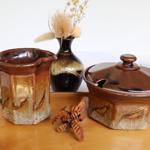 May include: A brown ceramic creamer and lidded sugar bowl set with a distinctive, geometric design. The set has a glossy finish with earthy tones of brown and tan. A small, dark vase with dried flowers and a wooden tray complete the still life.