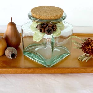 May include: A clear square glass jar with a round cork stopper, featuring a decorative cluster of small brown seed pods and pale green leaves attached to the neck. The jar is accompanied by a wooden apple, a wooden pear, and a dried flower.