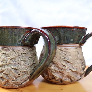 May include: Two ceramic mugs featuring a dark, glossy green glazed rim and handle, contrasted with a textured, matte beige lower body. The surface of the lower section has a rough, uneven, and organic appearance, while the upper rim is smooth.