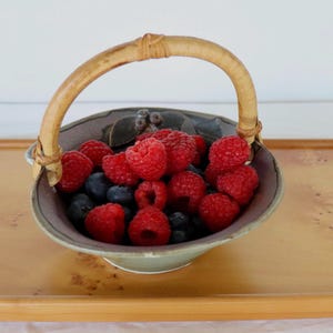 May include: A ceramic bowl with a curved light brown handle filled with fresh red raspberries and dark blue blueberries. The bowl features a dark grey interior with a small sculpted leaf and berry detail on the rim, resting on a light wood tray.